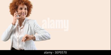 Smiling businesswoman with curly red hair talking on smartphone and checking time on watch, professional communication and time management Stock Photo