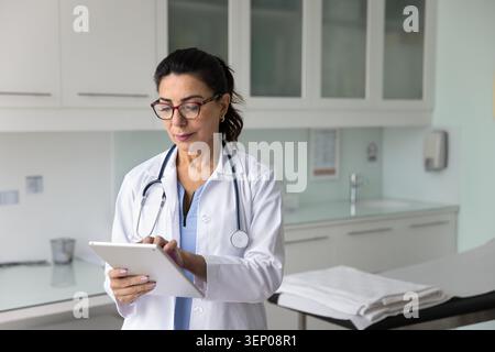 Serious senior Latin doctor wearing glasses, typing on tablet Stock Photo