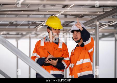 Two engineers inspecting solar panel infrastructure with digital tablet, discussing maintenance work under photovoltaic roof. engineering colleagues d Stock Photo