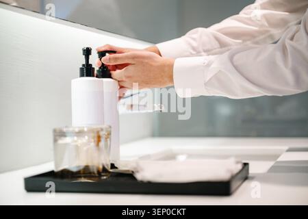 Hand presses white soap dispenser hand cleaning on counter in white restroom. Person wears a crisp white shirt, Glass jar sits nearby on black tray. C Stock Photo