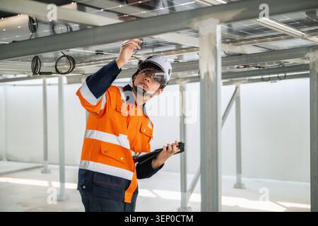 Young engineer in orange uniform and white helmet inspects solar panels, checking wiring cable under solar panels while holding a tablet check power c Stock Photo