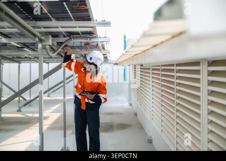 Young engineer in orange vest and helmet adjusts solar panel. Metal framework supports the panels. Industrial roof space with ventilation grilles. Sun Stock Photo