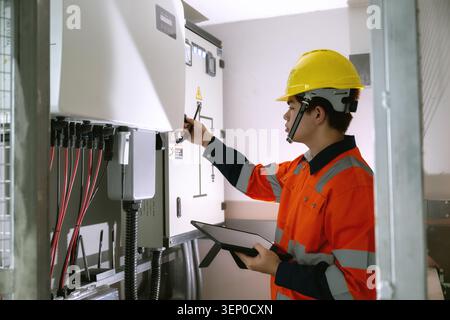 Technician in orange uniform maintaining control panel power station, holding tablet operating solar inverter switch, digital device inspecting solar Stock Photo