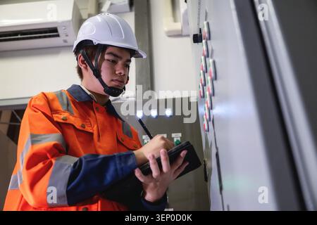 Asian young electrical engineer holding digital tablet inspecting main control panel board and writing data, Inspecting building switch panel factory Stock Photo