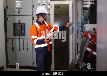 Asian young IT engineer in uniform using tablet and points to blue cables. Inspecting server rack network data center, maintaining telecommunication s Stock Photo