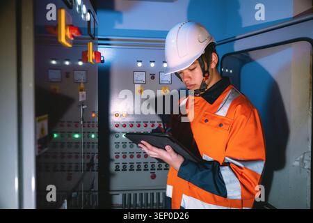 Young electrical engineer in orange safety clothing and a white helmet checking control panel. Control panel has buttons and green lights. Concentrate Stock Photo