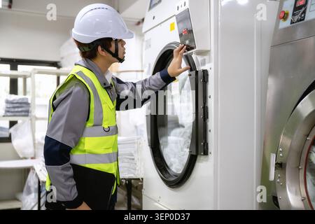 Maintenance technician pressing button on industrial laundry machine control operating large tumble dryer, adjusting settings near industrial washing Stock Photo