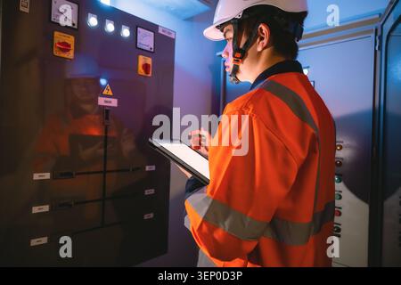 Worker in orange safety gear examines control panel. He wears a white hard hat and holds a clipboard. Bright lights illuminate warning signs and label Stock Photo