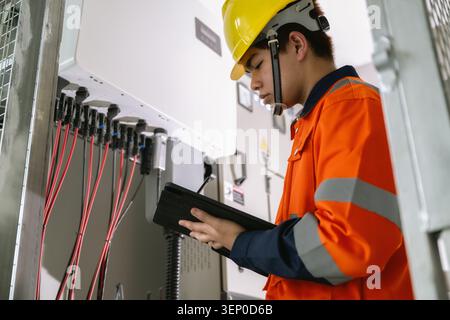 Young technician using tablet tracking solar power electrical circuit. wearing orange safety vest and yellow hard hat checks electrical panel with tab Stock Photo