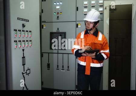 Asian young electrical engineer holding digital tablet inspecting main control panel board, young technician in orange uniform writing data near elect Stock Photo