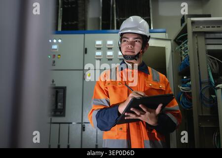 Asian young electrical engineer recording operation metrics on digital tablet screen indoor in control room, Electrician in a control room with equipm Stock Photo