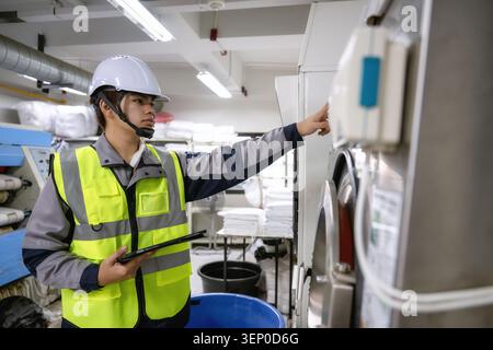 Maintenance technician holding tablet operating industrial laundry machine control, checking large washing machine factory subsystem, adjusting settin Stock Photo