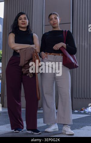 Diverse women wearing pants standing on sidewalk by concrete panels holding tan bag dark red tote Stock Photo