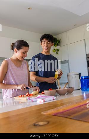 Diverse couple preparing fruit bowls at kitchen island, slicing strawberries and peeling banana Stock Photo