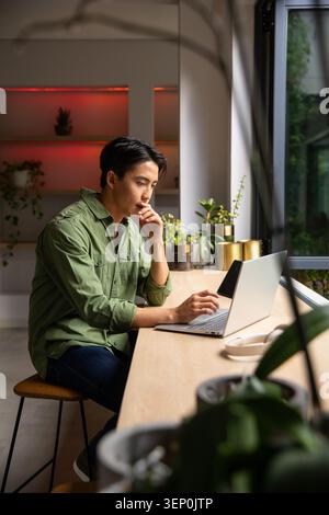 Asian adult man sitting, working on silver laptop with headphones and potted plants at wood counter Stock Photo