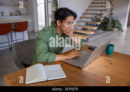 Asian man sitting at table at home wearing green shirt, typing on laptop, open notebook nearby Stock Photo