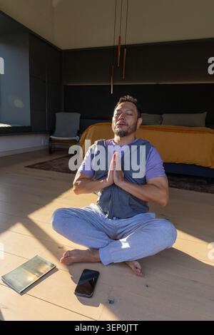 Asian man sitting cross-legged meditating on wood floor in bedroom, smartphone and notebook nearby Stock Photo