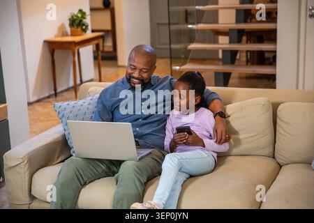 African American father blue-shirt and child sitting on beige sofa using laptop and phone at home Stock Photo