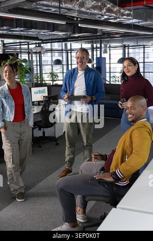 Diverse coworkers talking around senior man holding tablet in open-plan office, denim and mustard Stock Photo