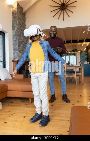 African American father and son playing in living room child wearing VR headset father using tablet Stock Photo