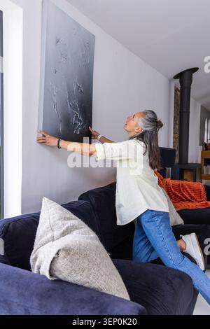 Senior woman standing on navy sofa straightening dark-gray canvas in living room, copy space Stock Photo
