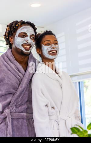 African American couple standing in home spa by window, wearing plush robes and clay masks Stock Photo