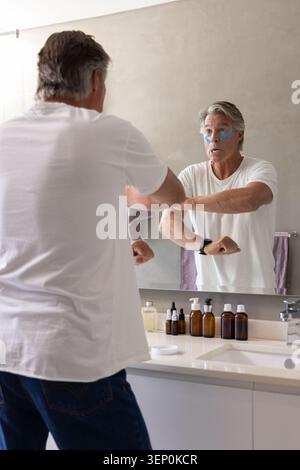 Senior man wearing white T-shirt and dark blue jeans applying under-eye patches at vanity mirror Stock Photo