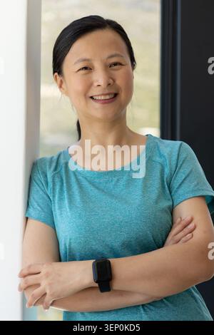 Asian woman leaning against white wall by large window, wearing teal shirt and black smartwatch Stock Photo