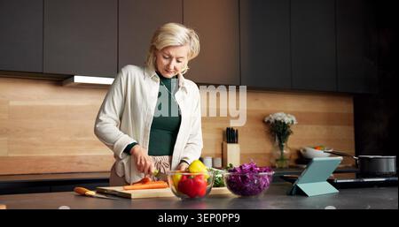 Woman, cooking and vegetables in kitchen with tablet, recipe and meal prep for healthy food. Mature person, digital and online video at house with Stock Photo