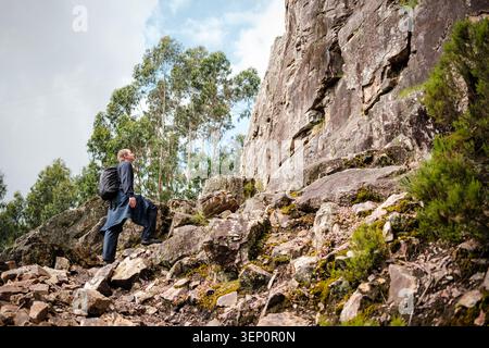 Man with backpack hiking a rocky mountain trail, looking up to the sky Stock Photo