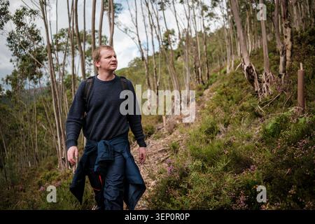 Man with backpack walking uphill on a trail through the woods Stock Photo