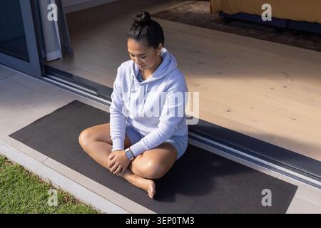 Asian woman sitting cross-legged on black mat in hoodie and shorts at door with smartwatch Stock Photo