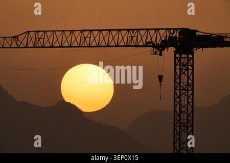 A glowing sun sets behind misty mountains, framed by the silhouette of a construction crane at dusk. Stock Photo