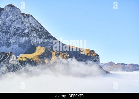 Jagged mountain cliffs emerge above a sea of fog, bathed in warm morning light and clear sky. Stock Photo