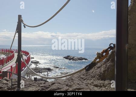Coastal scene showing the sea through protective nets, with rocks in the water, a small bridge on the side, and sunlight reflecting on the ocean Stock Photo