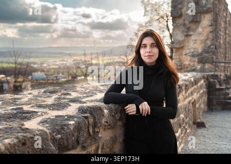 Young woman standing by an old stone wall in an outdoor historic setting with dramatic sky and scenic rural background Stock Photo