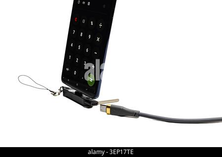 A black smartphone displaying a calculator app is held upright on a tiny portable USB-C docking station with a lanyard, positioned next to a disconnec Stock Photo