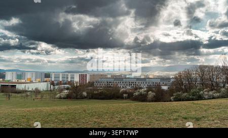Wide cityscape with apartment buildings, open field, distant hills, and dramatic storm clouds with sun rays breaking through the sky Stock Photo