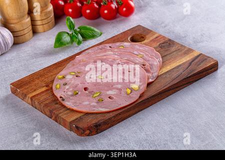 Thin slices of Italian mortadella with pistachios arranged on a rustic wooden board, surrounded by fresh basil, cherry tomatoes, garlic, and wooden mi Stock Photo
