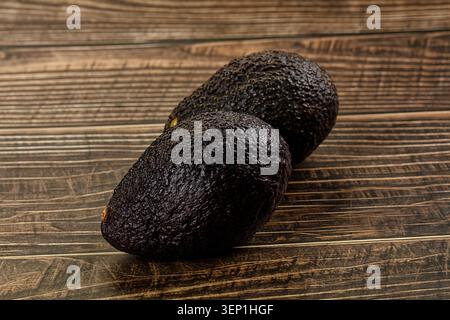 Two dark green avocados with bumpy skin resting on a textured wooden surface. Fresh organic fruit for healthy eating and cooking Stock Photo