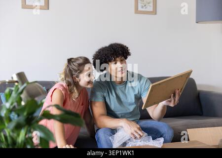 Couple sitting on gray sofa inspecting wooden frame with bubble wrap and box in living room Stock Photo