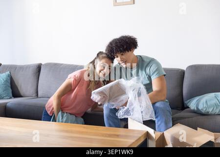 Couple sitting on grey sofa in casual clothes inspecting bubble-wrapped item by coffee table Stock Photo