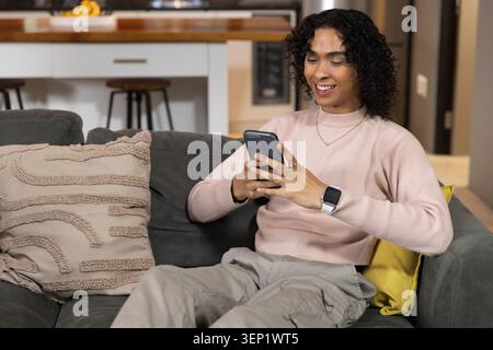 Asian man in his 20s sitting on gray sofa at home, using smartphone and smartwatch Stock Photo