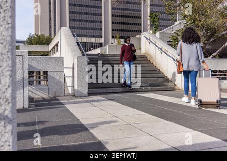 African American man descending stairs holding phone while woman pulling wheeled suitcase at plaza Stock Photo