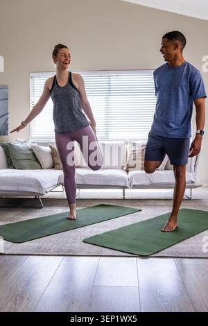 Diverse couple starting workout, doing quad stretch on mats at home wearing sportswear and trackers Stock Photo