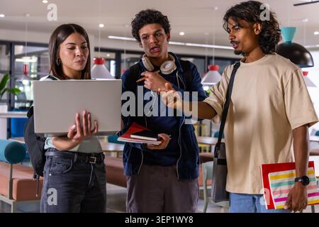 Diverse college students studying in study lounge around silver laptop, holding notebooks, pointing Stock Photo