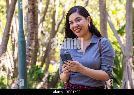 Adult woman smiling and holding phone with hands in shaded park with red plaid, brown bag Stock Photo