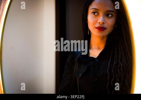 Young mixed race woman looking away in a bright reflection, highlighting her braided hair, red lips, and focused expression, exploring themes of beaut Stock Photo