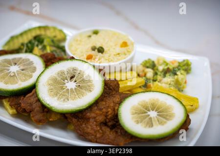 Delicious breaded steak meal served on a white plate. Featuring fresh lime slices. Seasoned rice. Mixed steamed vegetables. And golden french fries. R Stock Photo