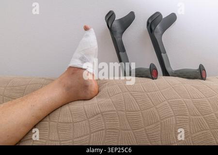 A bandaged foot after surgery to correct a bunion and two crutches leaning against the wall. Stock Photo
